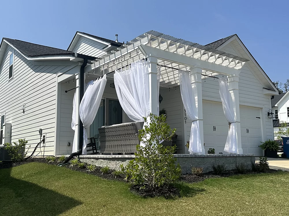 White pergola attached to home with flowing curtains, outdoor seating, and landscaped patio